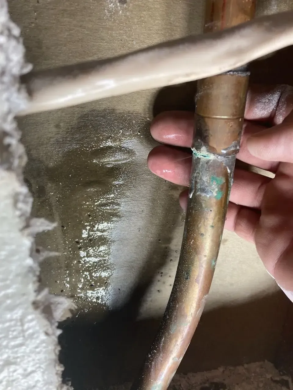 Hand touching a copper pipe with green corrosion, likely a leaky plumbing joint.