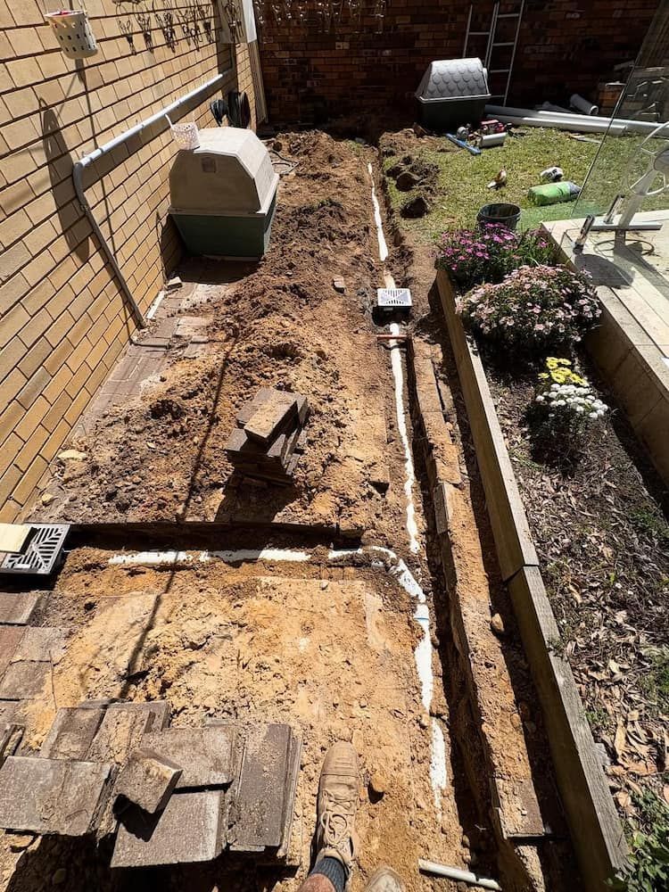 White PVC Drainage Pipes Are Laid in a Trench Beside a Brick House — Crystal Clear Plumbing & Roofing in Wollongong, NSW