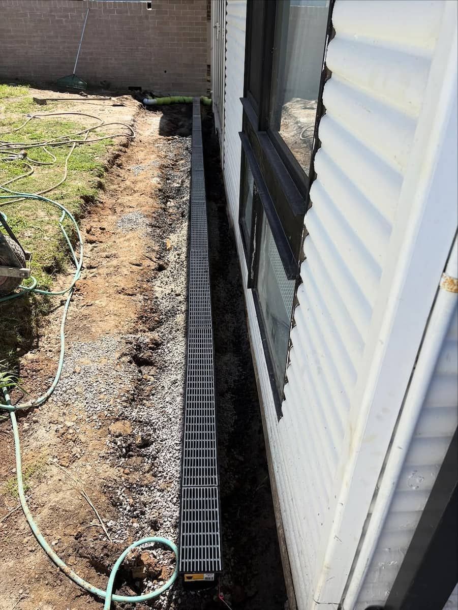 A Black Plastic Trench Drain Installed Along the Side of a White House — Crystal Clear Plumbing & Roofing in Austinmer, NSW