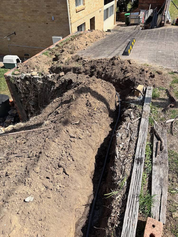 A Narrow Trench Dug Into a Dirt Yard, Showing Exposed Black Conduit — Crystal Clear Plumbing & Roofing in Austinmer, NSW