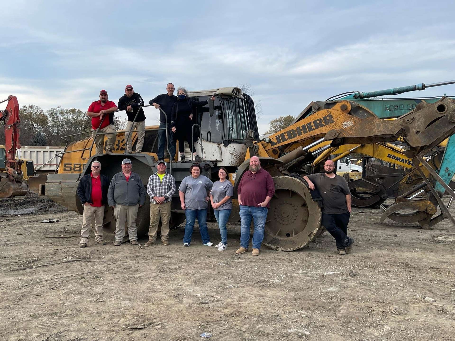 Group of people posing with heavy machinery at an industrial site on an overcast day.