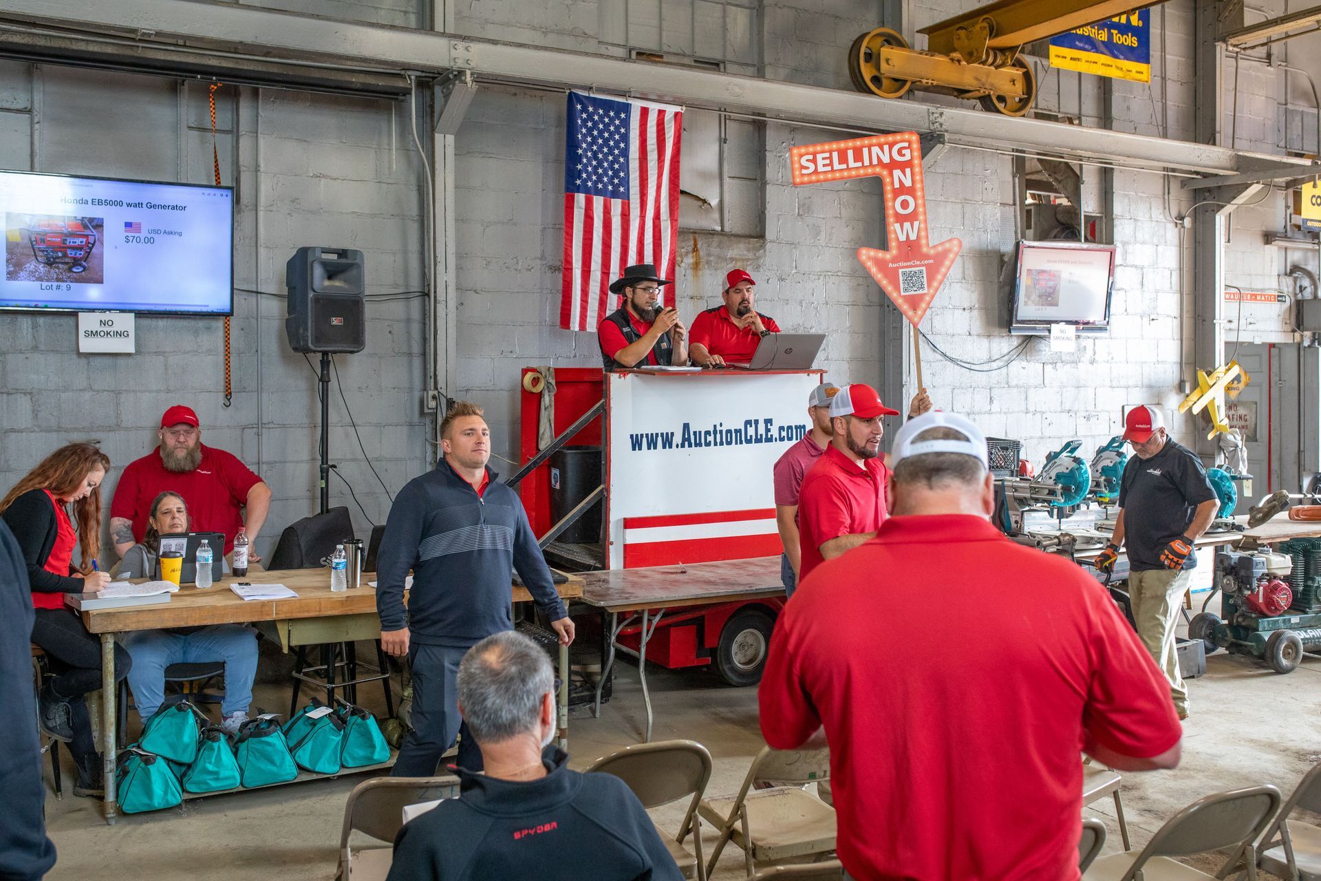Auction in a warehouse; people bidding, auctioneer at a podium with American flag.