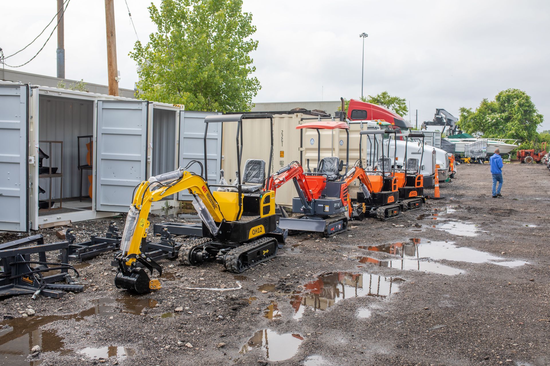 Row of excavators in a muddy lot near storage containers, a person walks by.