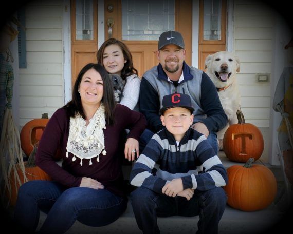 Family and dog sitting on porch steps with pumpkins. Smiling, wearing fall attire.