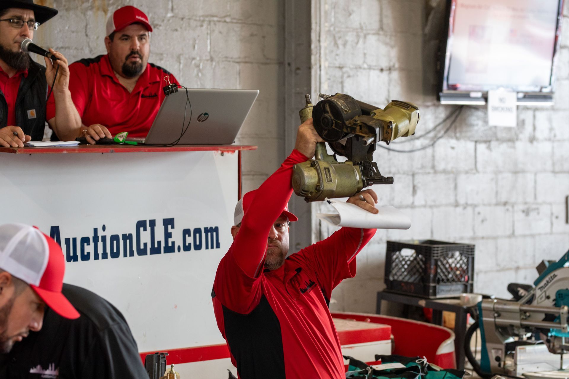 Auctioneer holding a mechanical part, two men at a table with laptop, inside a building.