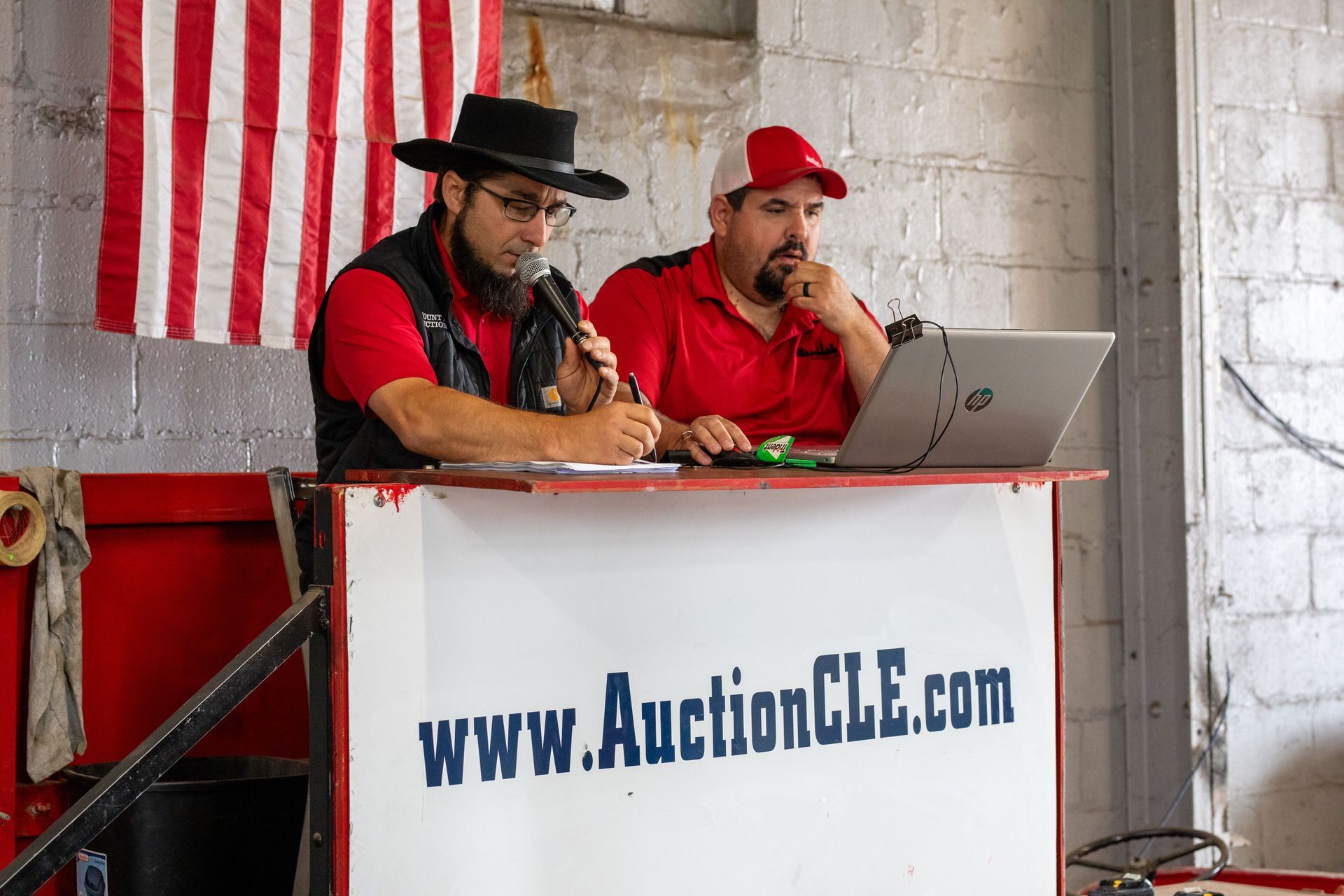 Two men at an auction. One speaks into a microphone, the other works on a laptop. An American flag hangs behind them.