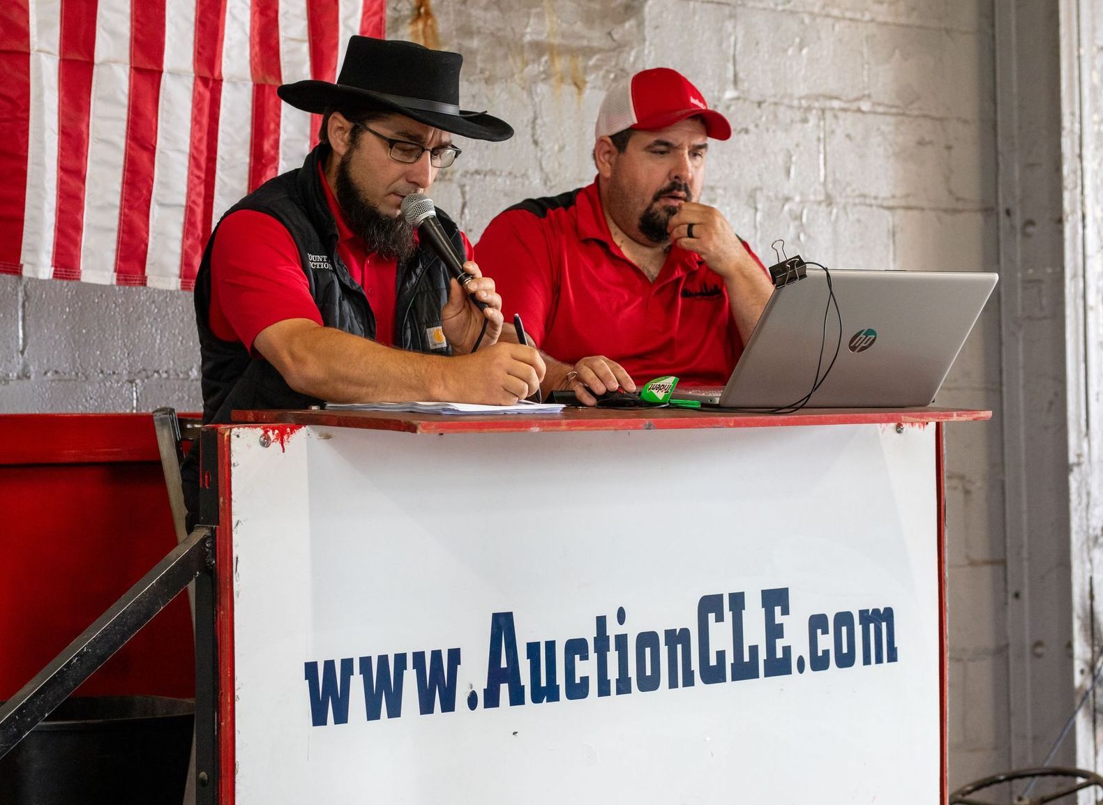 Two men at a red and white auction stand. One man holds a microphone and writes, other looks at a laptop.
