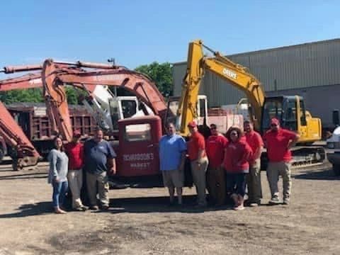 Group of people in red shirts and khaki pants stand in front of excavators and trucks.