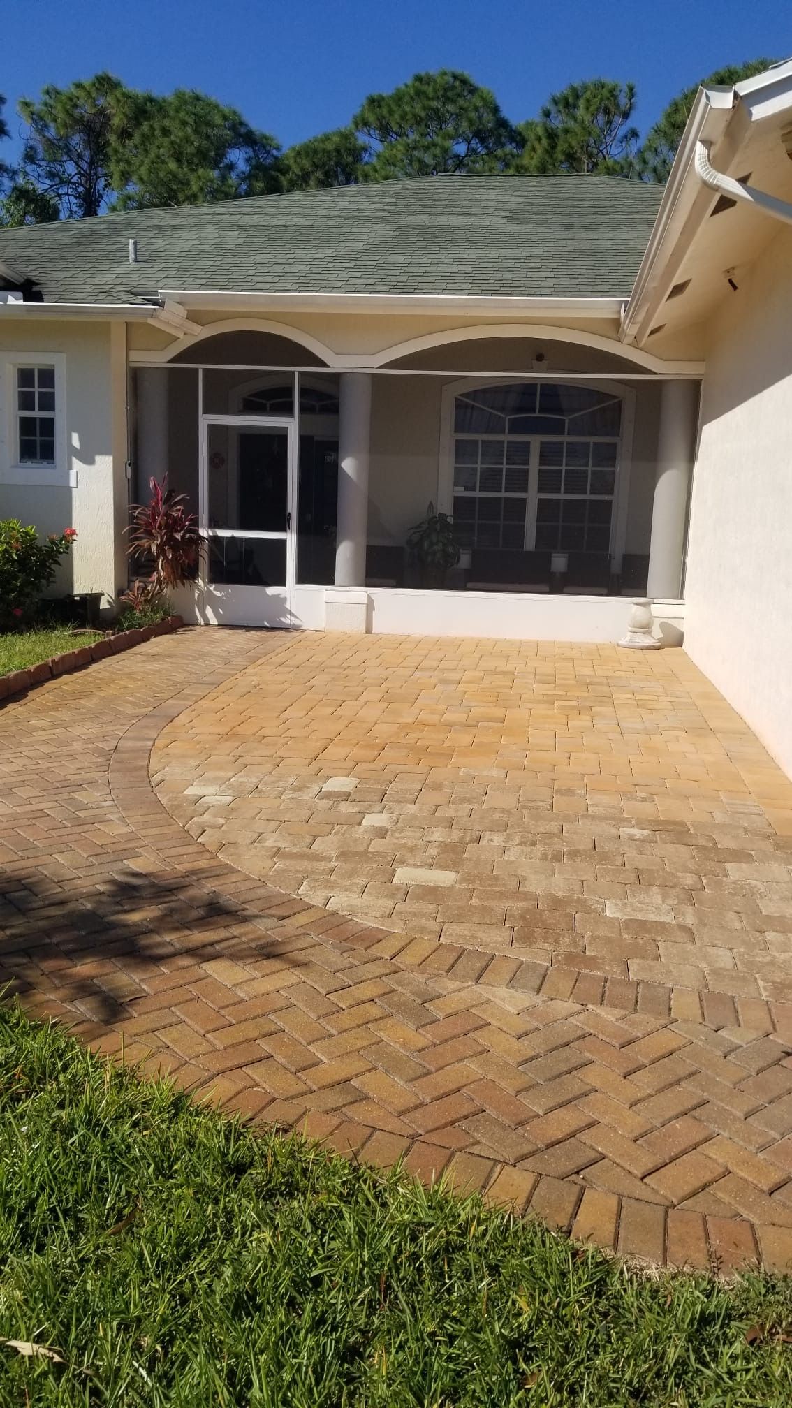 Brick paved patio leading to a screened porch with arched openings, surrounded by grass and a beige house.