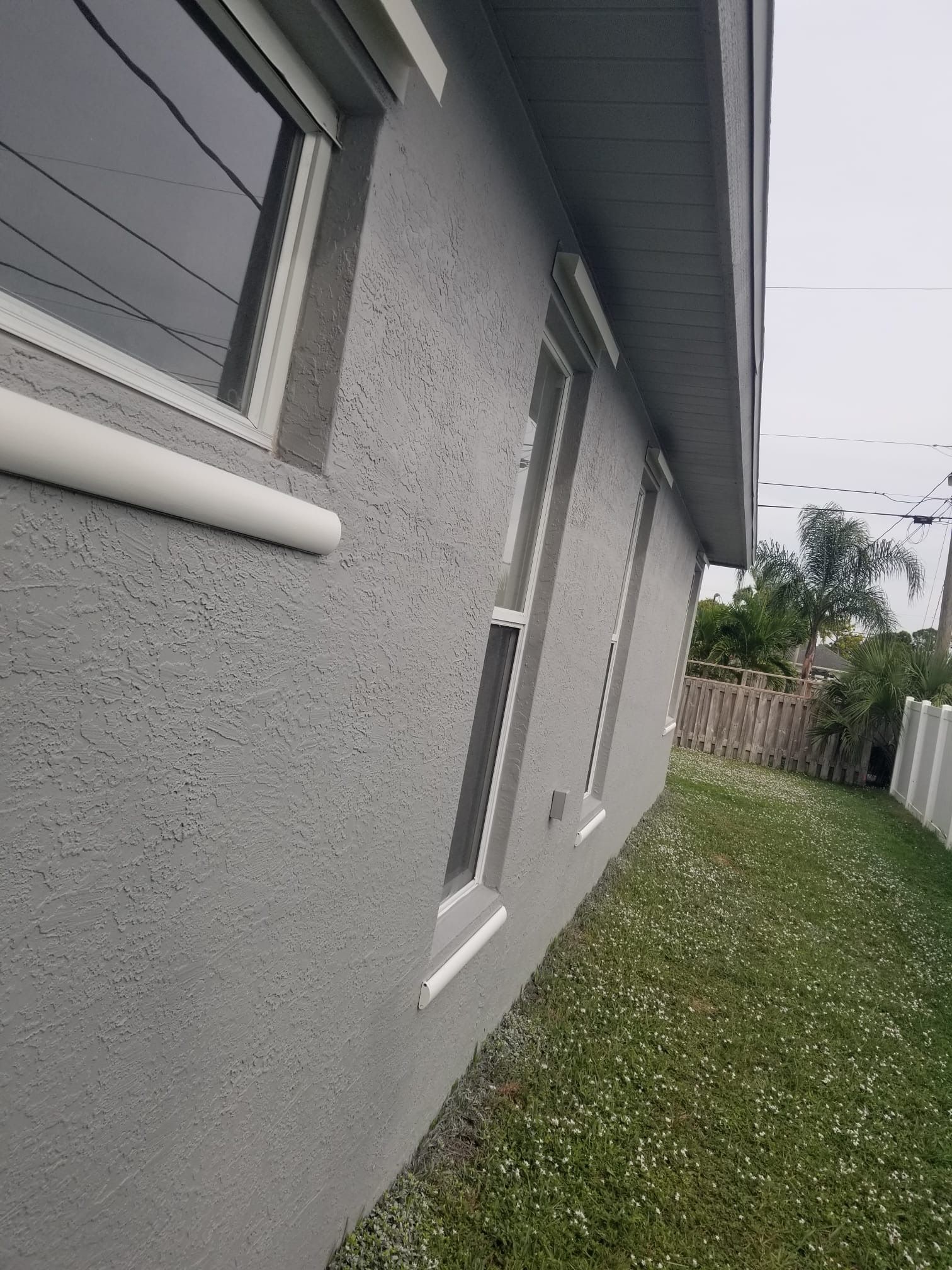 Gray stucco house exterior with windows, white trim, and a green lawn.