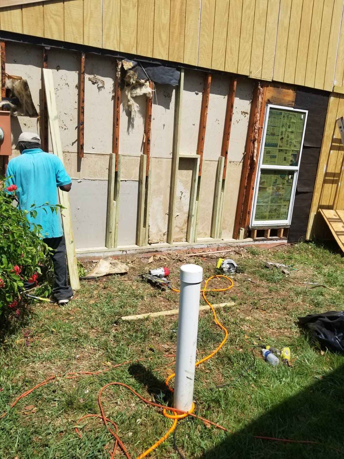 Man standing near a house wall undergoing repair, with exposed studs and construction tools on the grass.