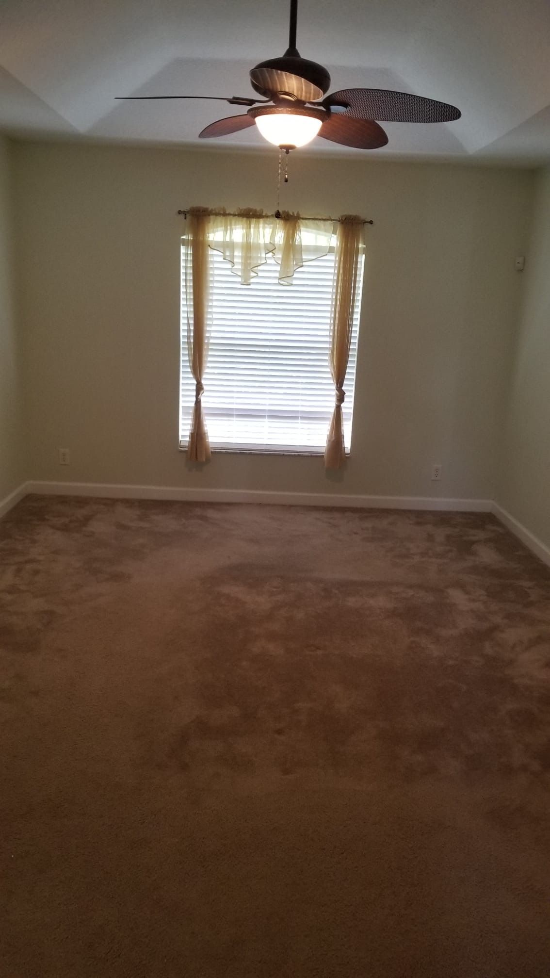 Empty bedroom with tan carpet, a window, and a ceiling fan.
