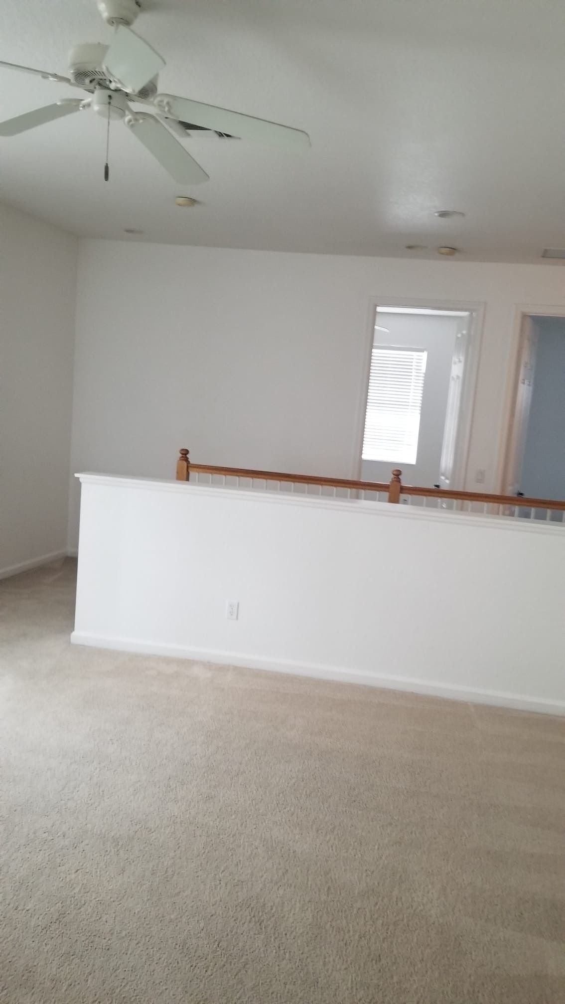 An empty room with a white wall and tan carpet. A ceiling fan and railing are visible.