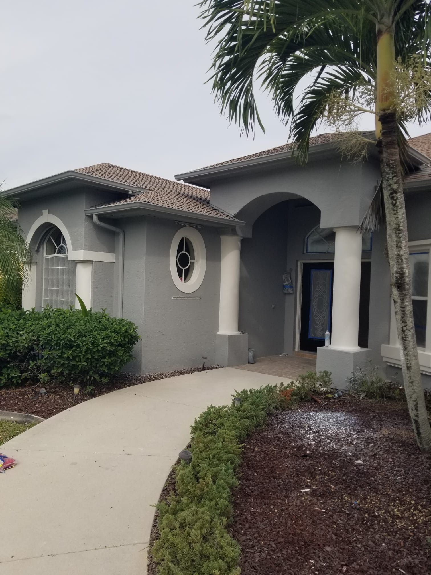 Gray stucco house with arched doorway, white columns, and palm tree. Sidewalk curves toward entrance.