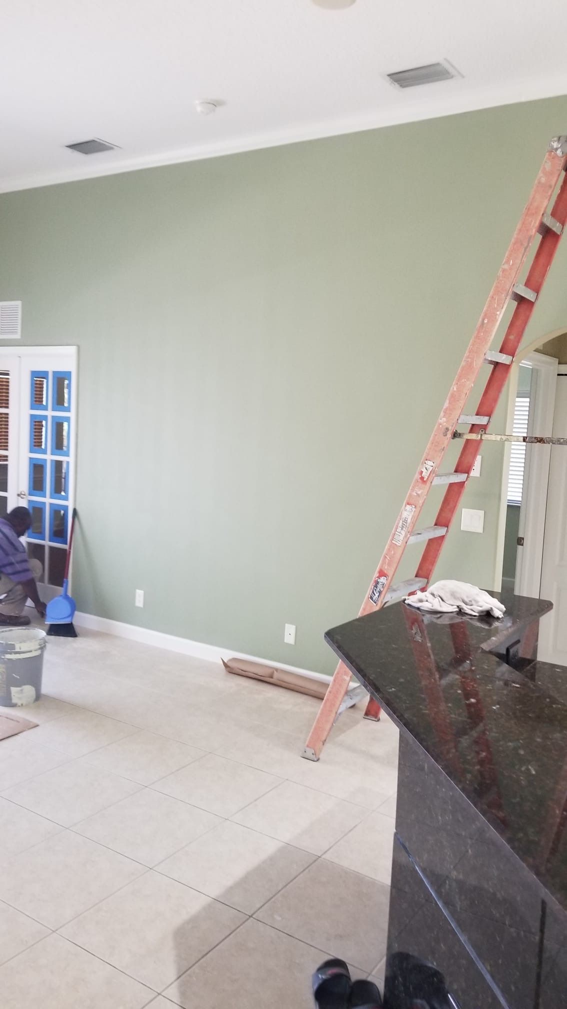 Person painting green wall, orange ladder, open door, kitchen counter, tiled floor.