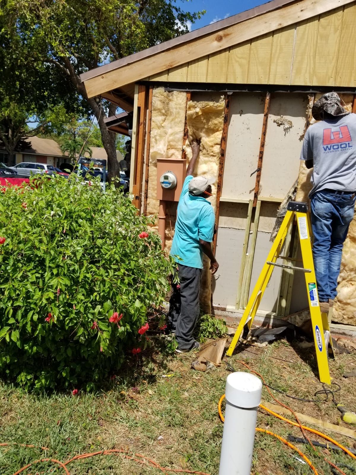 Two people removing exterior siding from a house. One is reaching up, the other is on a ladder.