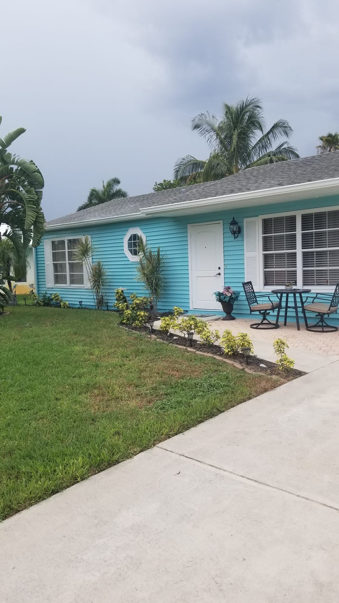 Turquoise house with white trim, door, and windows. Green lawn, sidewalk, and cloudy sky.