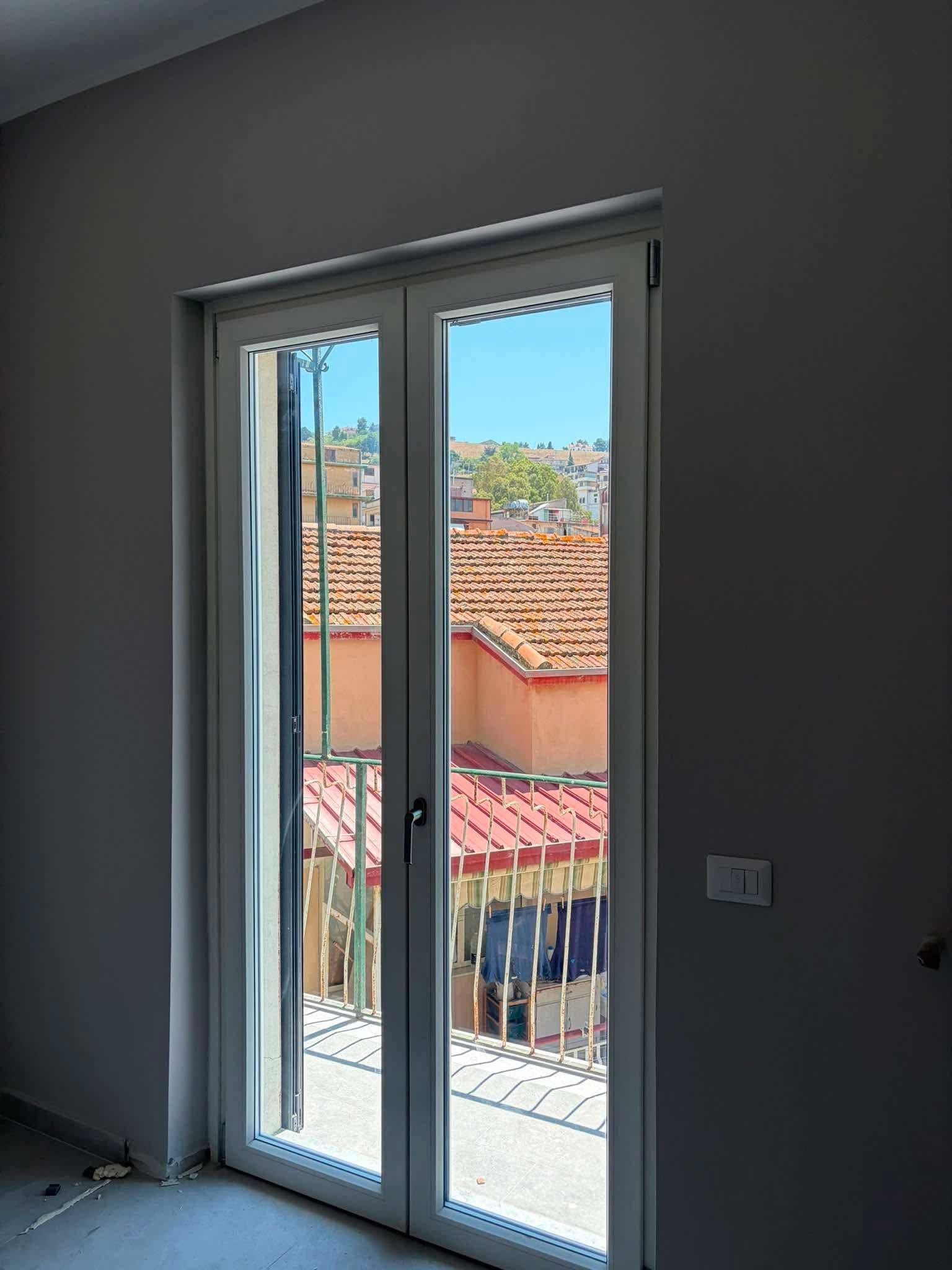 Vista dall’interno di porte-finestre bianche che si aprono su un balcone, con tetti di tegole rosse all’esterno