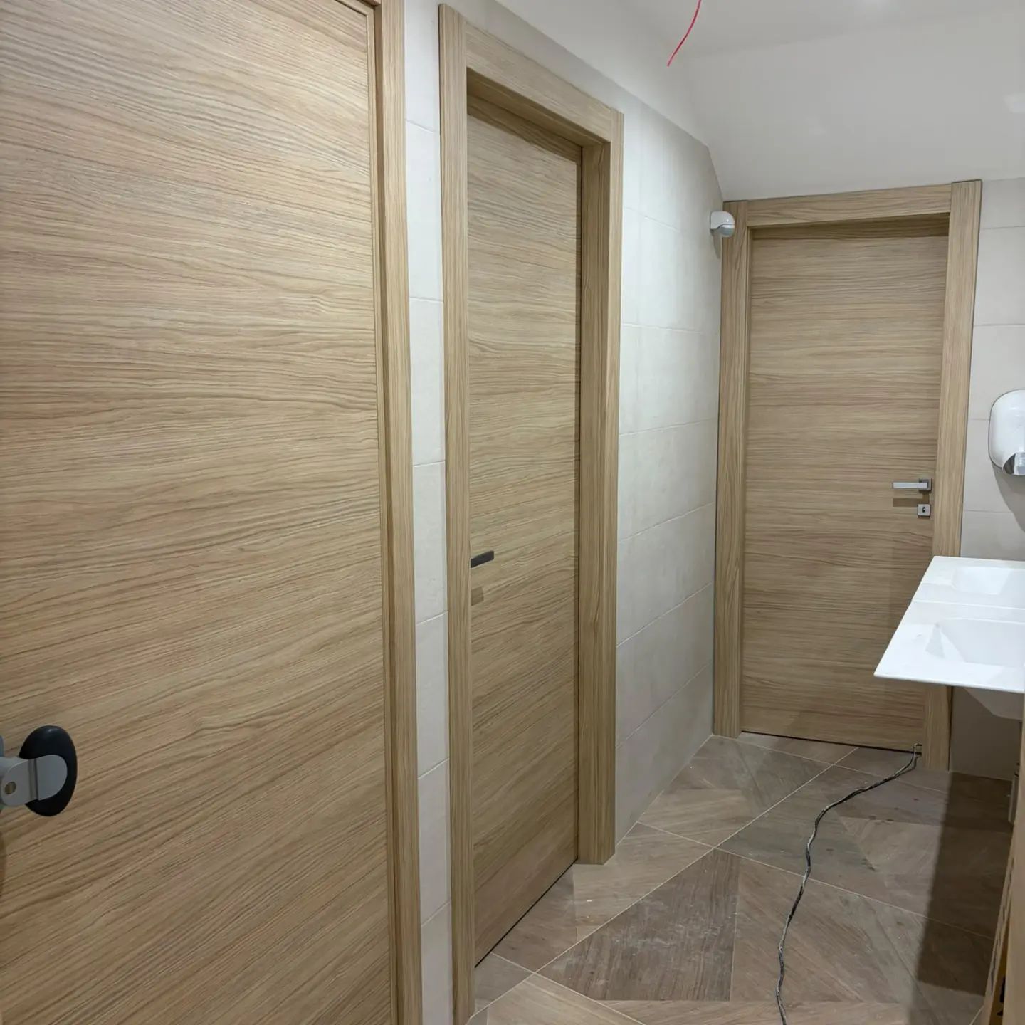 Corridoio stretto con porte in legno chiaro e pavimento piastrellato beige, in un interno moderno.