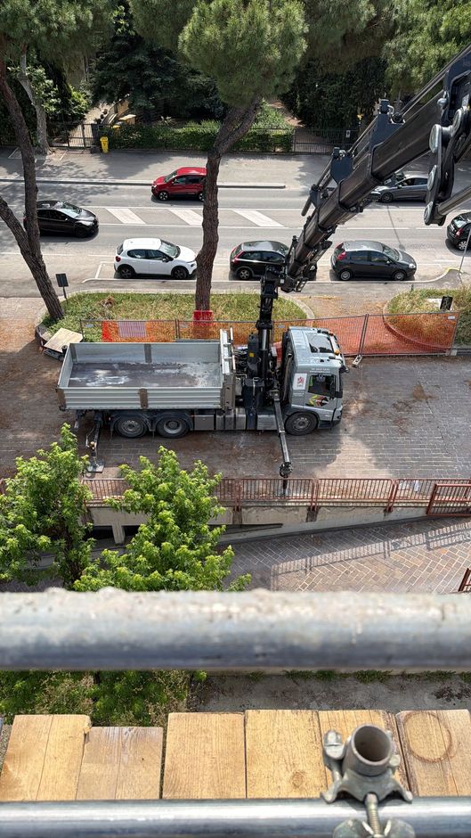 Camion con gru sollevata che carica rottami metallici in un piazzale sterrato.