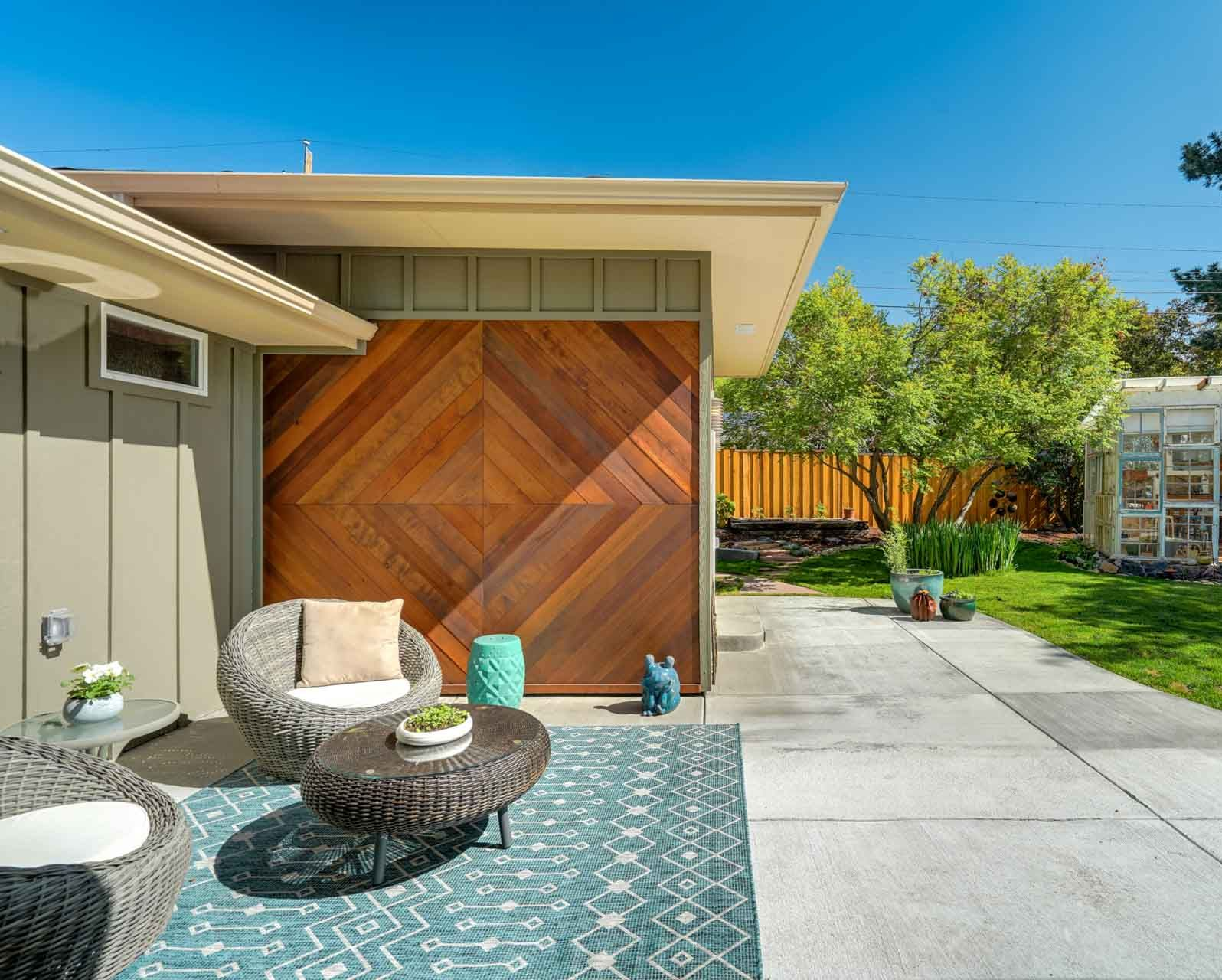Patio with wicker chair, patterned rug, and wood-slat wall beside a sunny concrete walkway and garden