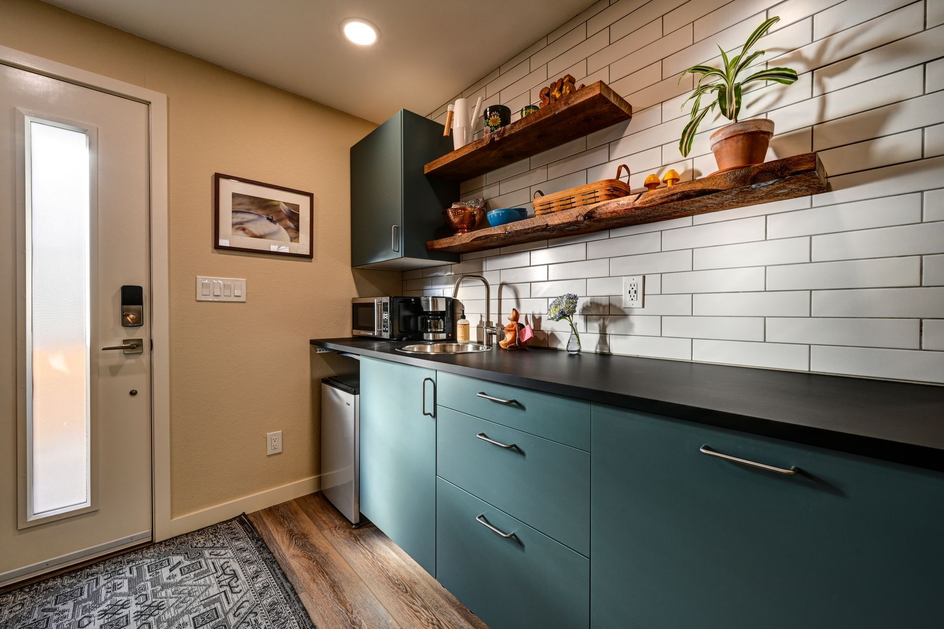 Modern kitchen with teal cabinets, white subway tile backsplash, wood shelves, and a door on the left.