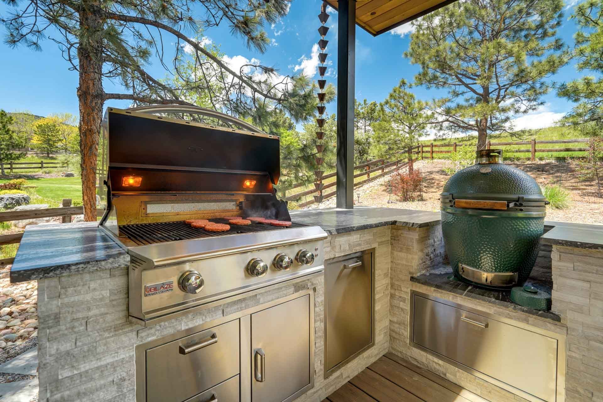Outdoor stainless steel grill on a stone counter under a patio cover, with a green ceramic smoker nearby.