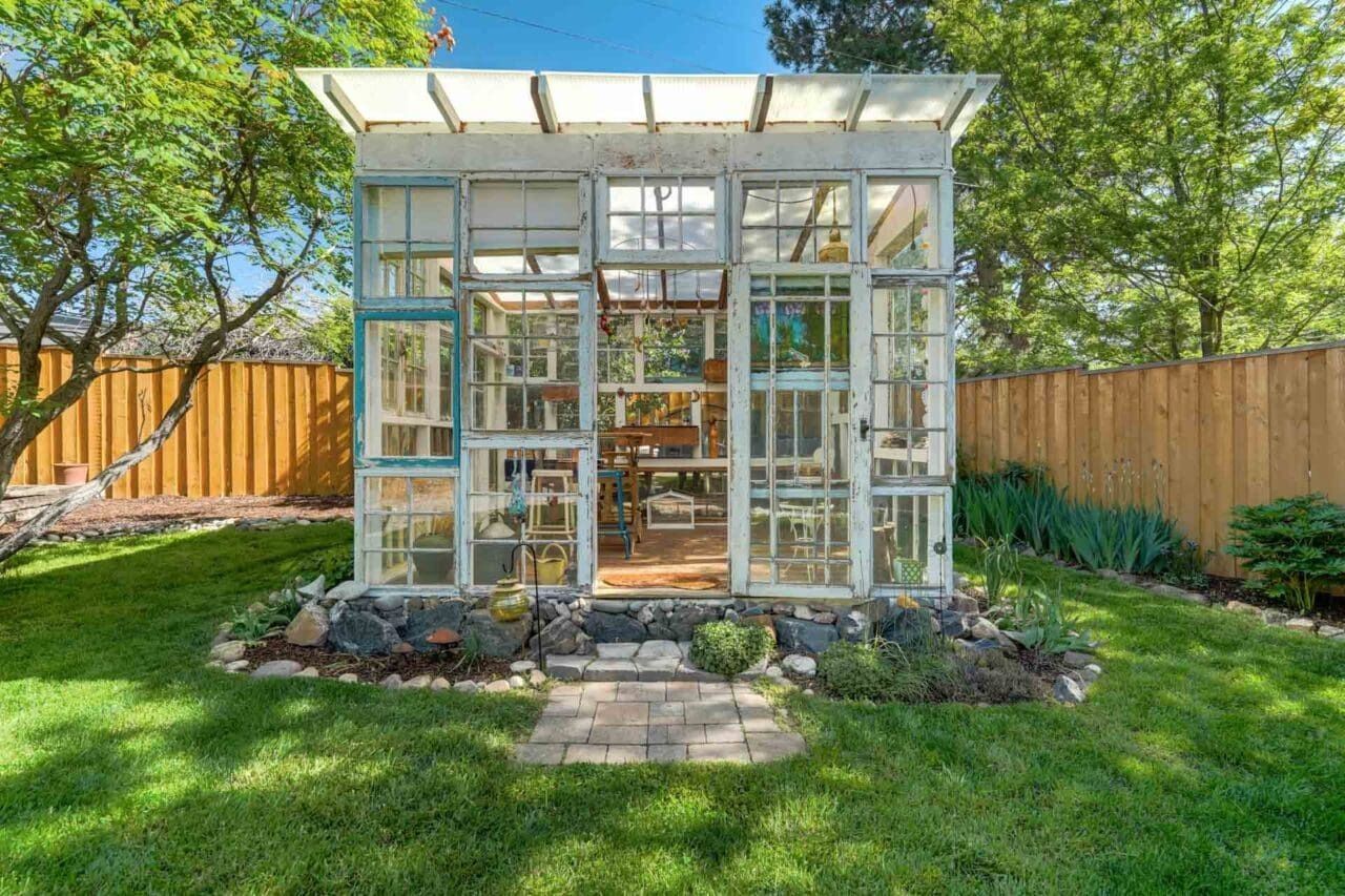 Glass greenhouse in a sunny backyard, framed by a wooden fence and lush green lawn.