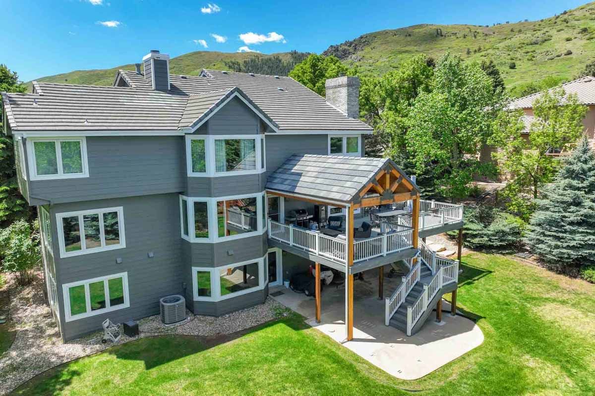 Gray multi-story house with a covered deck and staircase on a green hillside under a blue sky