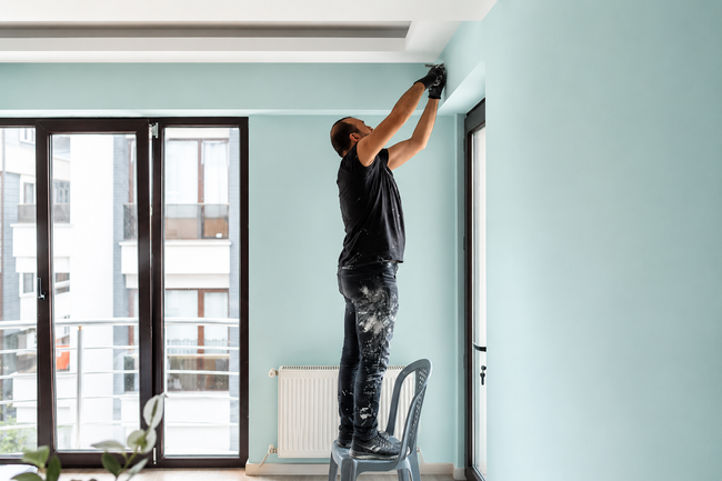 Person painting a light blue wall in a bright room, standing on a chair and using a roller.