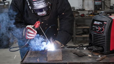 Welder in black protective gear using a torch on metal, with bright sparks and smoke in a workshop