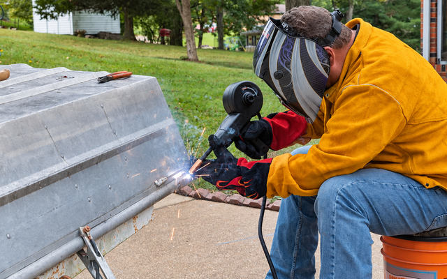Worker in a yellow jacket welding a metal barrier beside a sidewalk, with sparks flying.