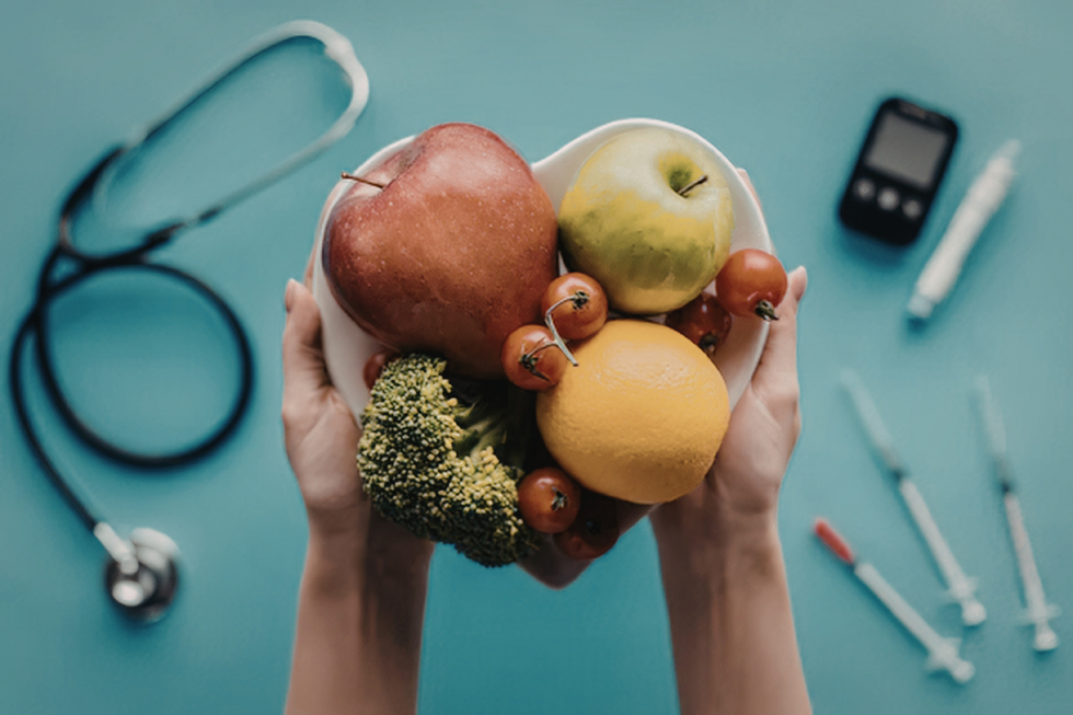 Hands holding assorted fruits and vegetables on a teal medical table with stethoscope and syringes