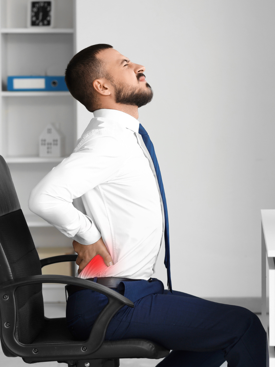 Man seated at a desk holding his lower back, grimacing in pain while wearing office attire