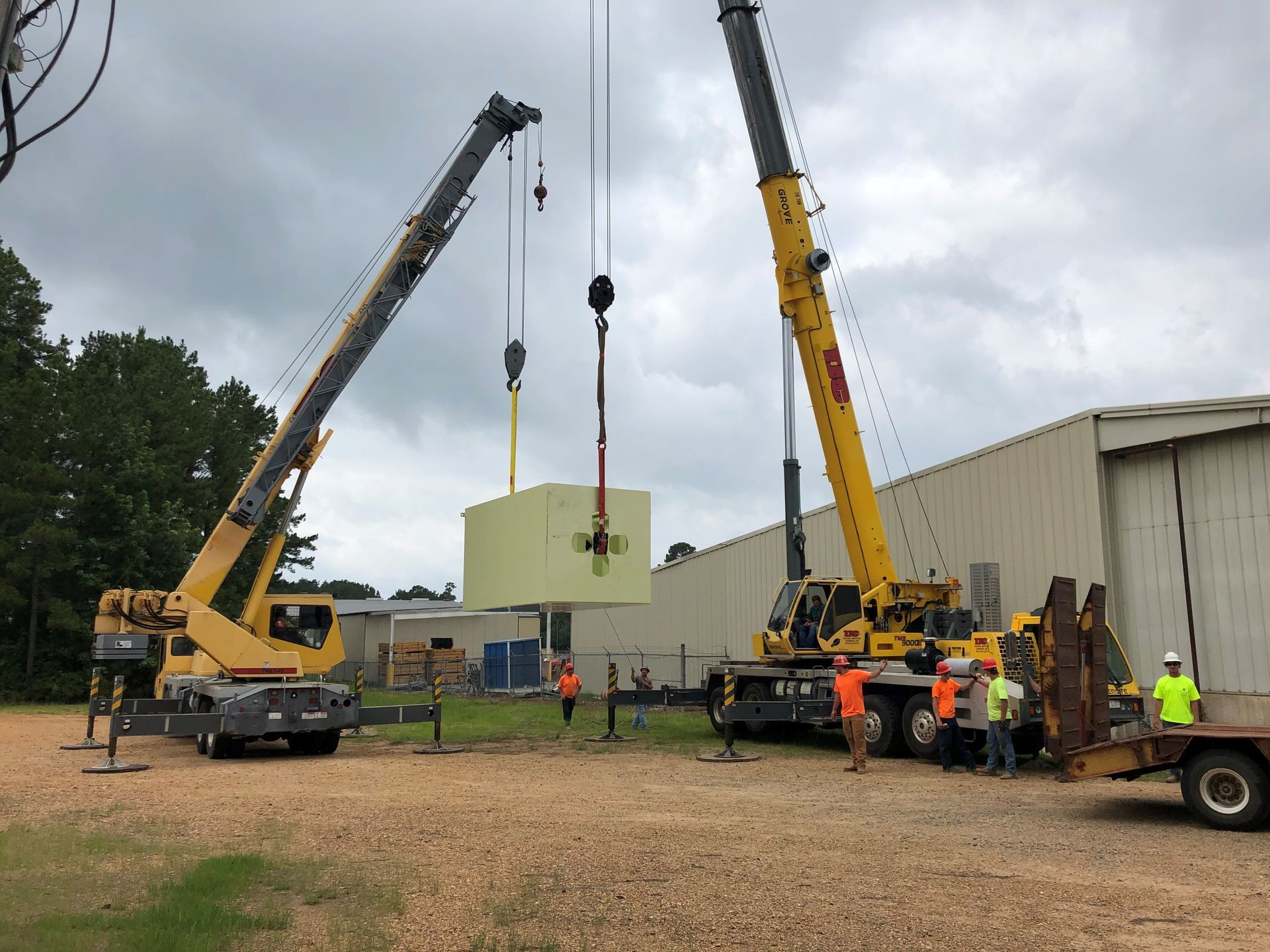 Two cranes lifting a large green tank beside a warehouse on an overcast day.