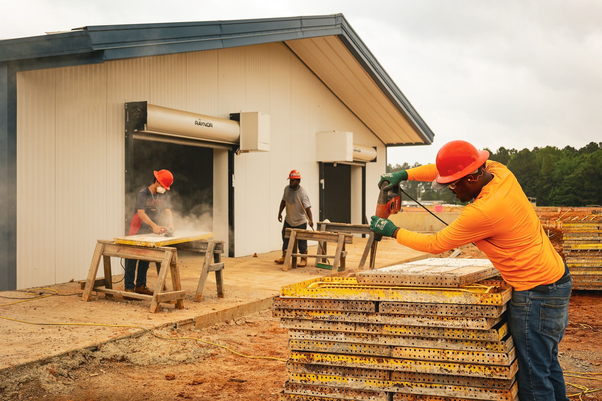 Construction workers laying roof shingles beside a small building on a muddy job site.