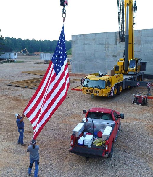 Two workers hold a large U.S. flag beside a red pickup and construction equipment on a gravel site