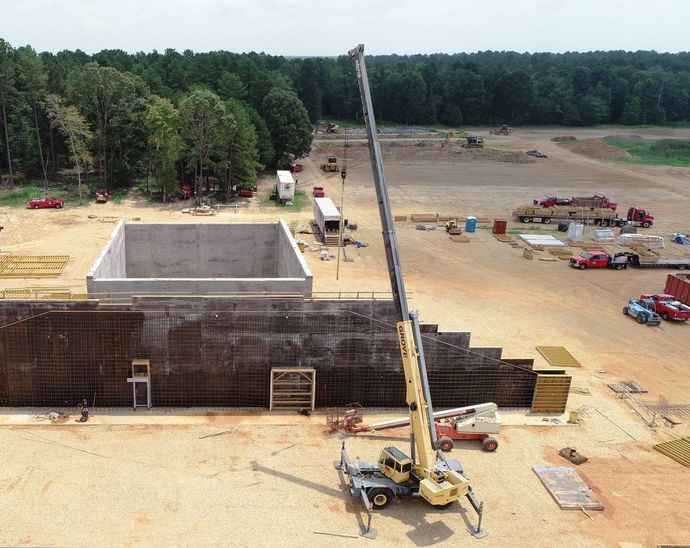 Construction site with concrete structure, crane, and trucks on a sandy lot near trees