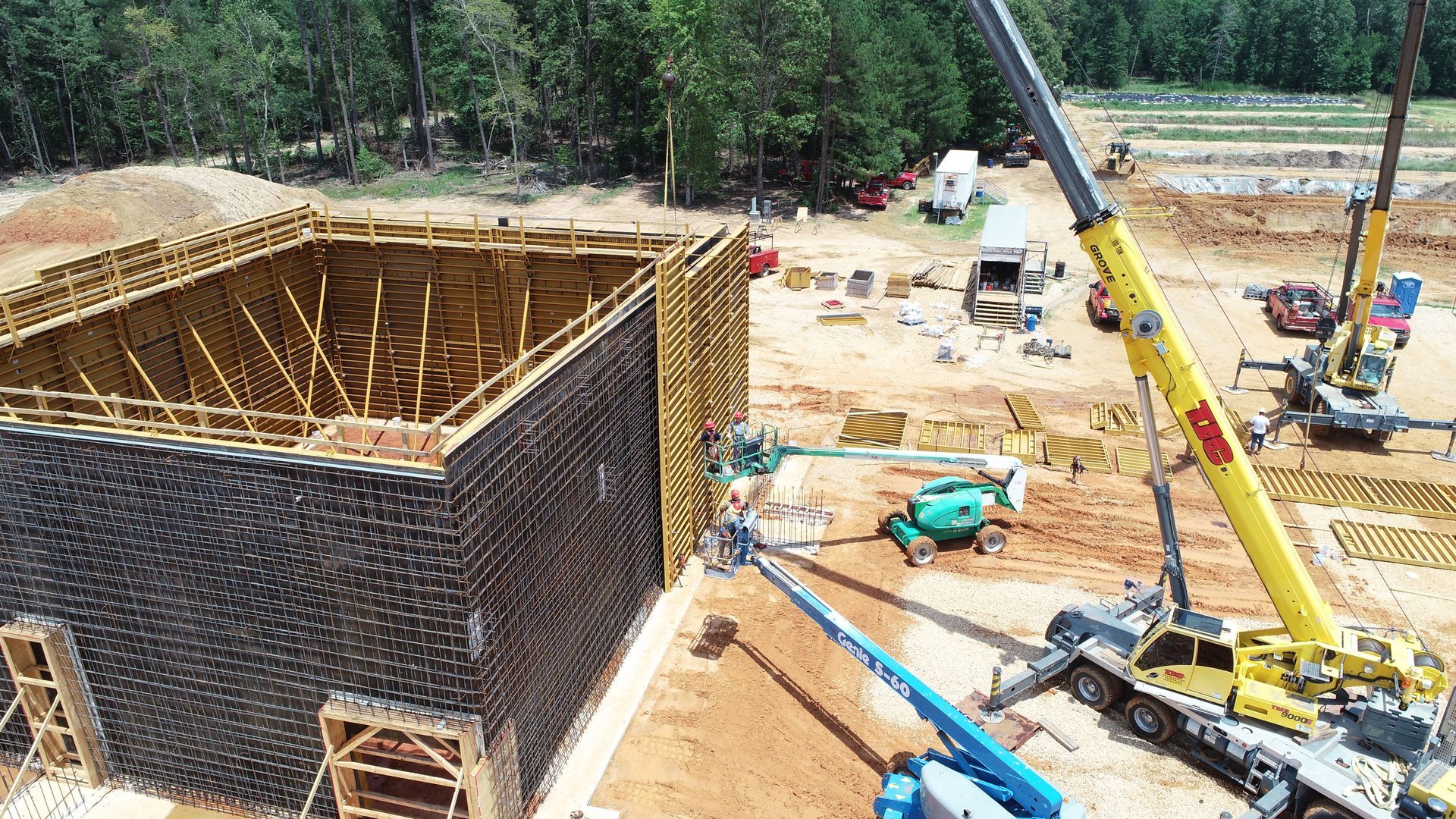 Construction site with brick building, cranes, workers, and heavy equipment amid wooded surroundings