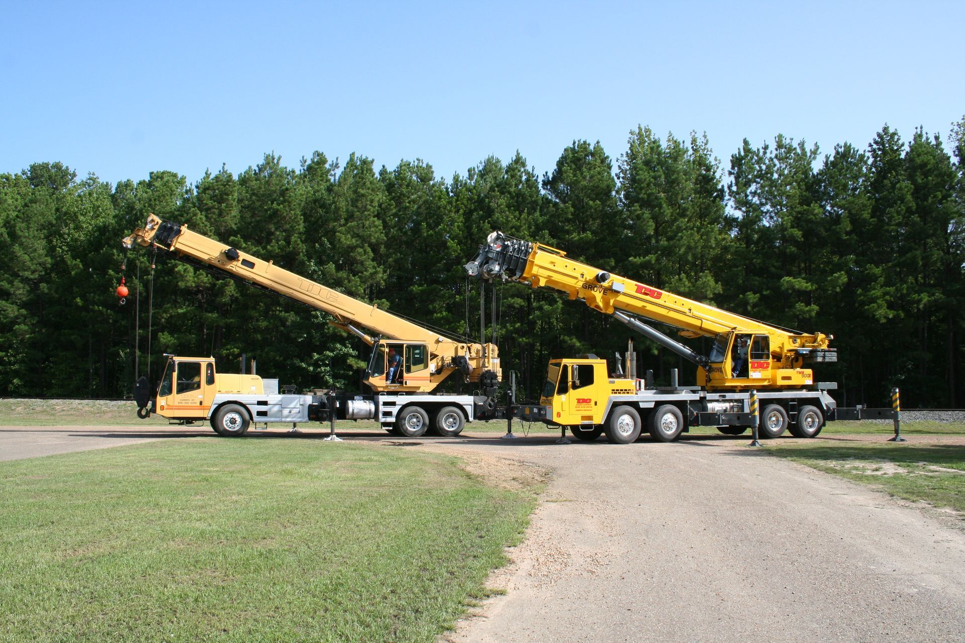 Two yellow utility cranes on trucks parked on a gravel lot beside a grassy field and trees