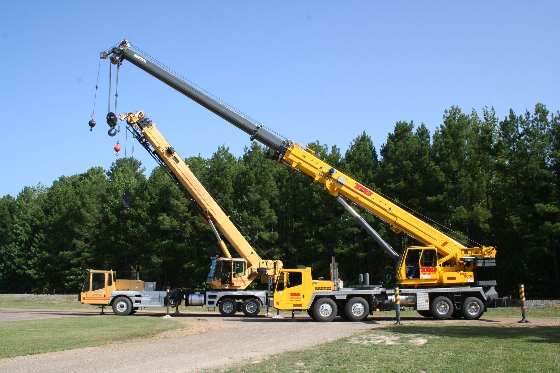 Two yellow construction cranes with extended booms on trucks in a grassy roadside area under a blue sky