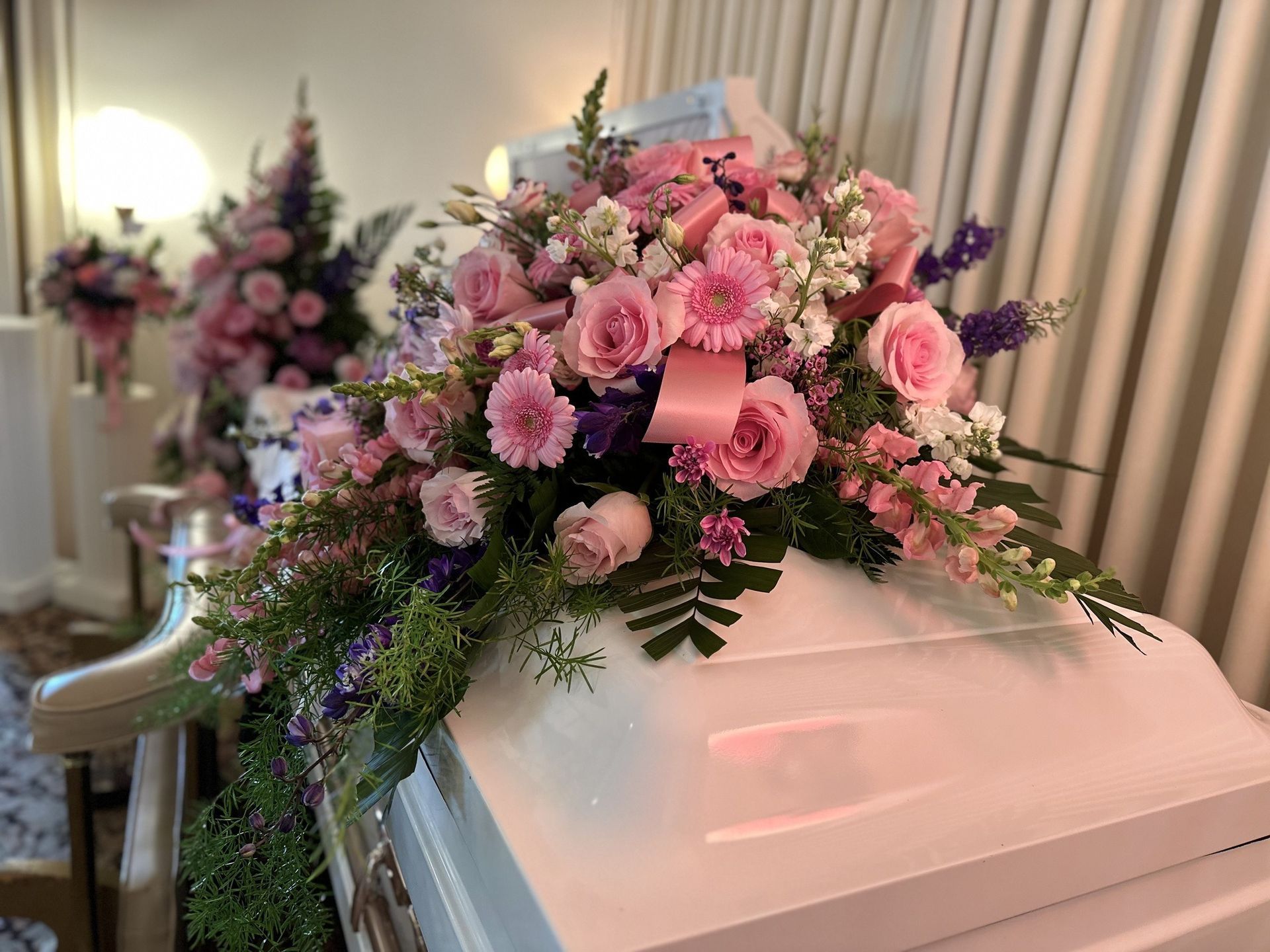 White casket adorned with pink flowers, in a room with more floral arrangements.