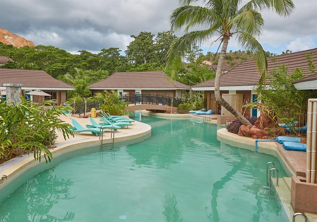 Tropical resort pool with turquoise water, palm trees, lounge chairs, and thatched-roof cabanas.