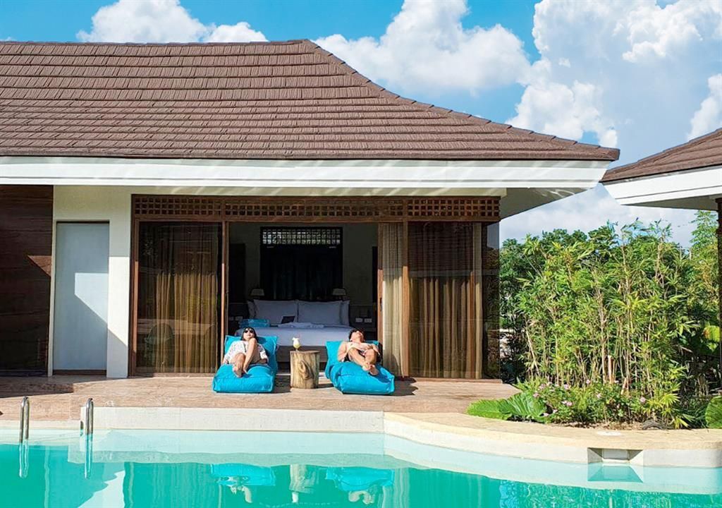 Two people in blue float in a pool before a tropical villa with open patio and greenery