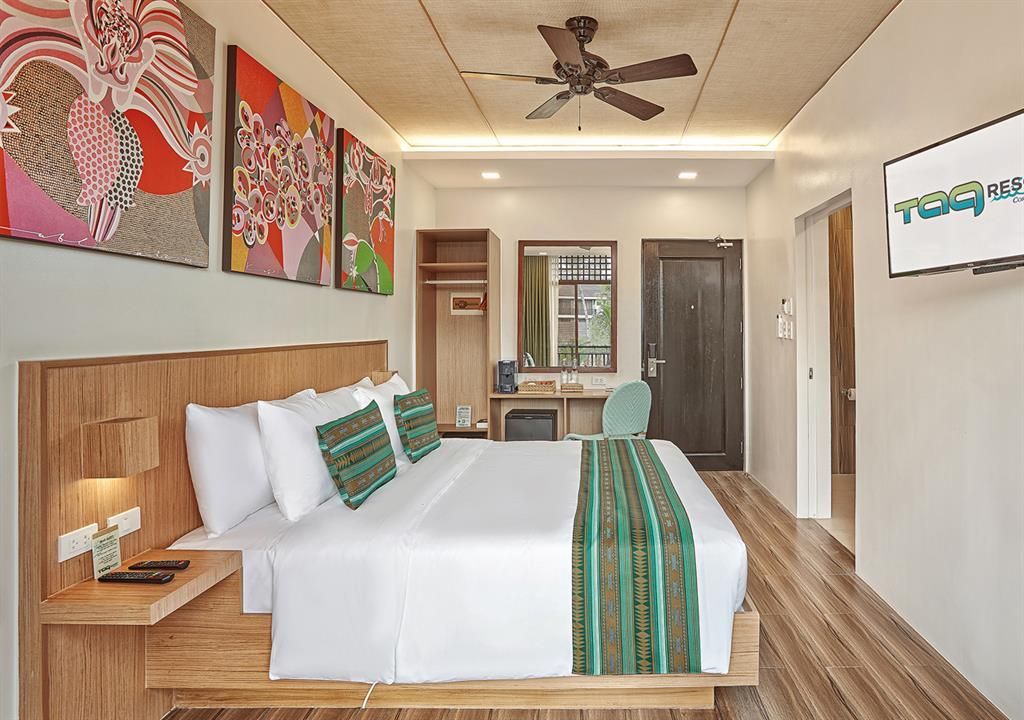 Modern hotel bedroom with a king bed, wood accents, wall art, ceiling fan, and balcony view