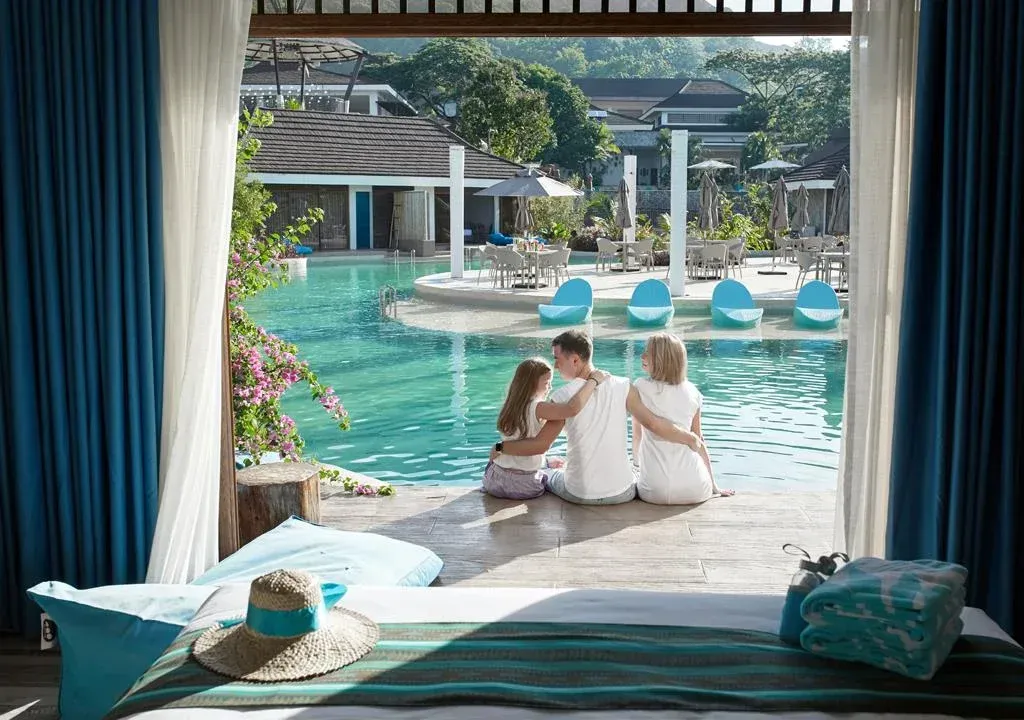 Two people sit by a turquoise resort pool, viewed from a shaded cabana with a bed in the foreground.