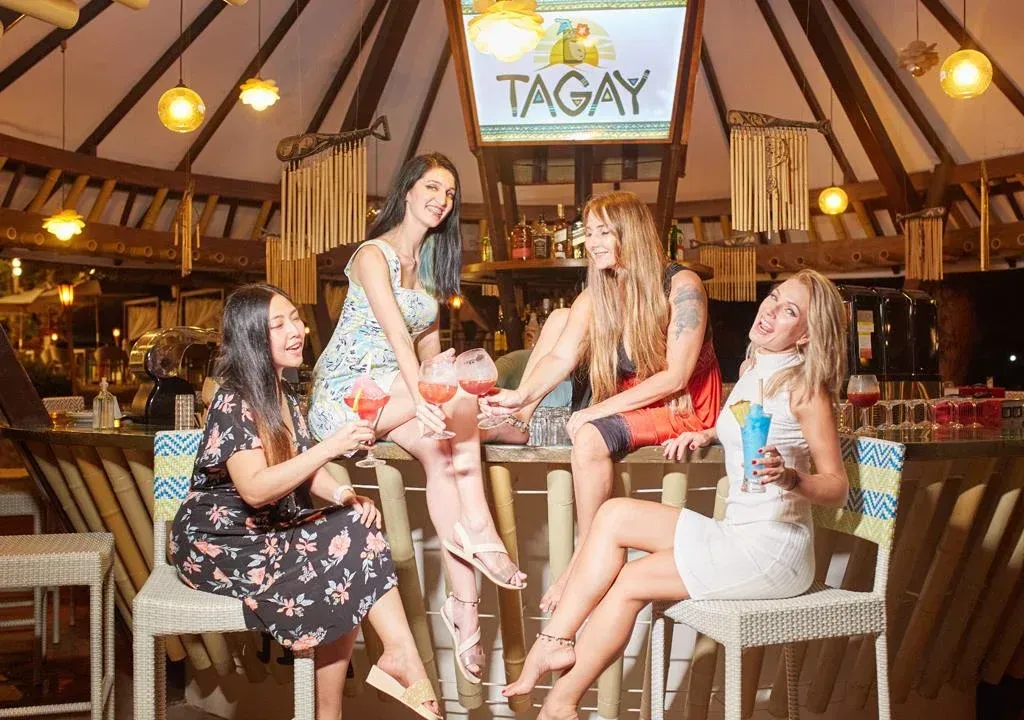 Five women chatting with drinks at a rustic indoor bar under a TAGAY sign