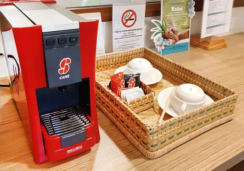 Red coffee machine beside woven tray with cups, saucers, and condiment packets on a wooden counter