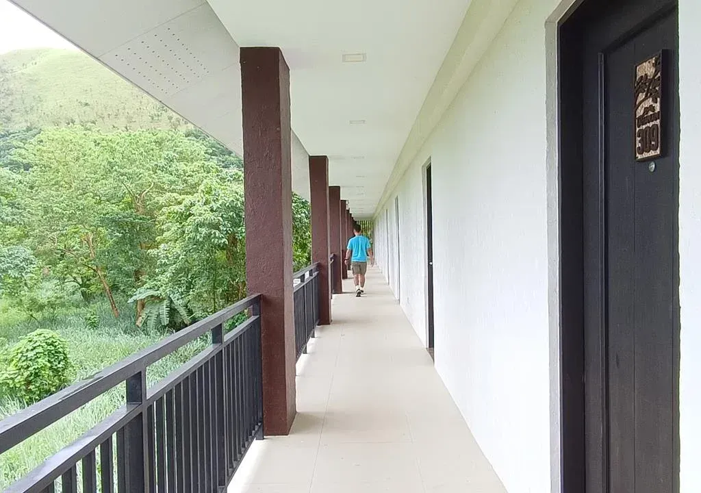Covered outdoor corridor beside a green hillside, with black railing and brown columns.