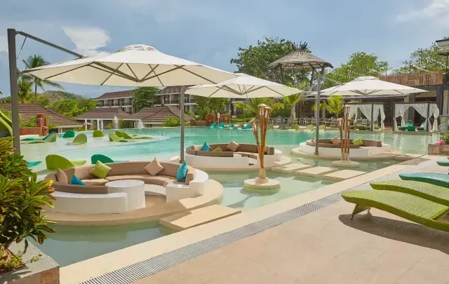 Sunlit resort pool with white umbrellas, lounge chairs, and turquoise water beside a tropical garden